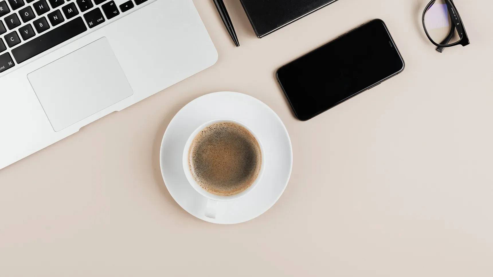 A laptop, coffee cup, phone and glasses on a light desk, seen from above