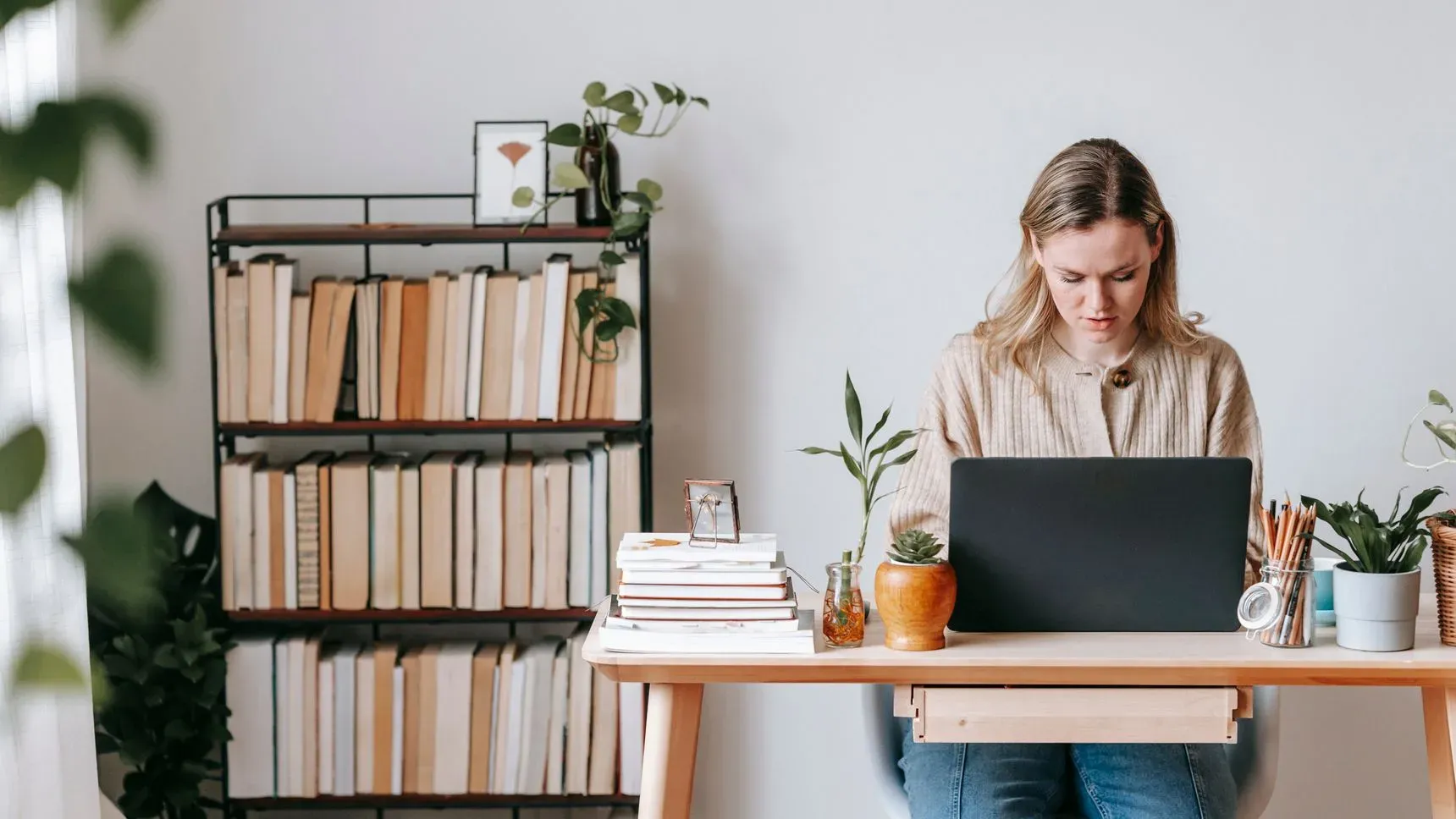 A woman working at a laptop at a desk with plants and a bookshelf