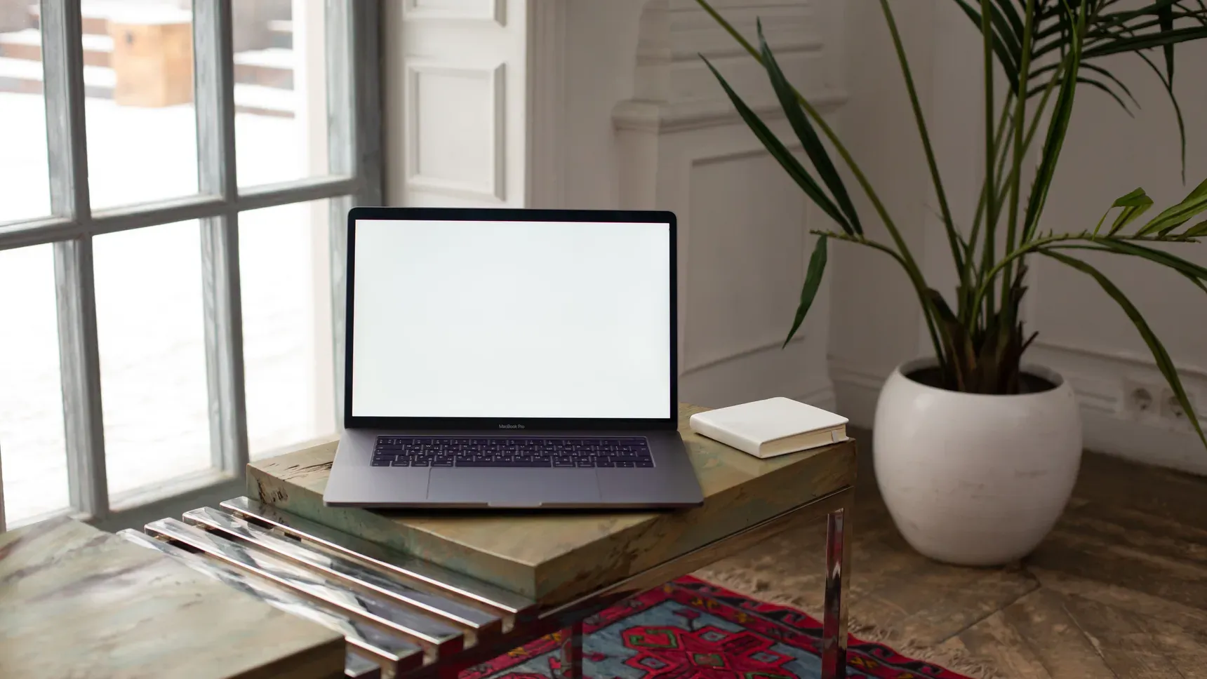 A laptop on a desk by a window with daylight and a plant